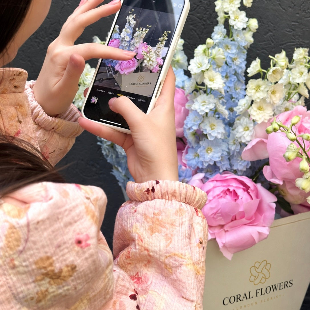 Coral Flowers bouquet of pink peonies and blue and white delphiniums in a branded box, being photographed on a phone
